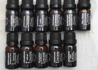 Flat lay of small amber glass essential oil bottles with black caps and black labels are arranged in two rows on a white textured fabric. Visible scent labels include Lavender, Lemongrass, Patchouli, Peppermint, Rosemary, Tea Tree, Bergamot, Cedarwood, Eucalyptus, Frankincense, and Grapefruit. Apothecuryous
