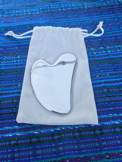 Silver Gua Sha tool on a white drawstring bag against a blue textured background