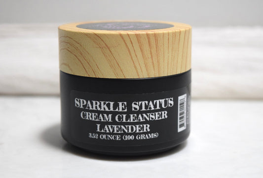 A jar of Sparkle Status Cream Cleanser in Lavender, featuring a black container with white text and a light wood-grain lid, set against a neutral gray background.